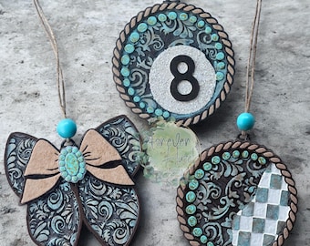 May include: Three hanging ornaments featuring dark brown surfaces with embossed turquoise floral patterns. One is a bow with a turquoise center bead, one is a circle with the number 8, and one is a circle with a white and turquoise checkered section.