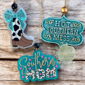 May include: Three hanging ornaments featuring a cowboy boot with a teal glitter bow, a teal plaque with the text HOT SOUTHERN MESS, and a teal sign with the text Southern Mom in gold glitter with leopard print and small pink hearts.