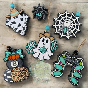 May include: A collection of six Halloween-themed ornaments featuring a cow-print ghost with a tan hat, a black spider, a spiderweb with a spider, a ghost with a tan hat and turquoise accents, an 8-ball pumpkin, and a turquoise-studded horseshoe.