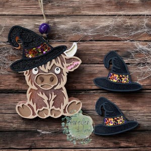 May include: A brown Highland cow cutout wearing a black glittery witch hat with colorful sequins, accompanied by two separate black glittery witch hats with colorful sequins. The cow has a light tan muzzle and white eyes with black pupils.
