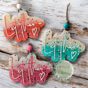 May include: Three cloud-shaped hanging ornaments with glittery gradients. One is orange-to-white with white text reading "let them", one is teal-to-white with white text reading "let them", and one is pink-to-white with white text reading "let them".