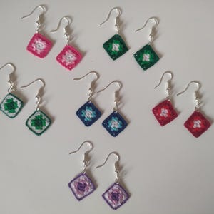 May include: A set of 10 crocheted earrings with silver hooks. The earrings are in various colours, including pink, green, blue, red, and purple. Each earring features a square-shaped crocheted design with a flower in the centre.