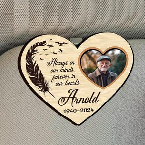 May include: A wooden heart-shaped memorial with a photo, a feather design, and the inscription "Always on our minds, forever in our hearts" and "Arnold 1940-2024". It is attached to a car sun visor.