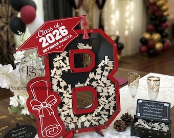 May include: A red and black graduation guest book alternative shaped like the number 8, filled with wooden scroll notes. Features a red mortarboard, diploma, and a sign reading "Class of 2026". Includes champagne flutes.