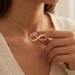 May include: A gold-colored infinity necklace with the names "Susan" and "Mark" engraved on it, along with the date "05.10.2022". The necklace is worn around the neck.