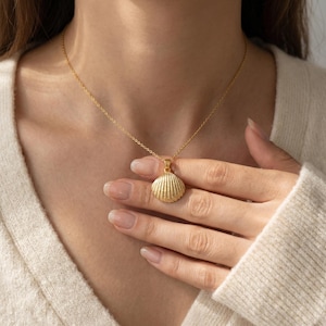 May include: A gold-colored necklace featuring a seashell pendant. The pendant is intricately detailed with ridged lines, and the chain is delicate. The necklace is displayed on a person wearing a cream-colored sweater.