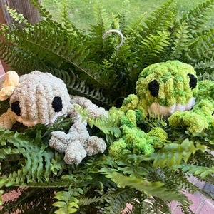 May include: Two crocheted plush toys shaped like frogs, one green and one grey, are nestled in a bed of green ferns.