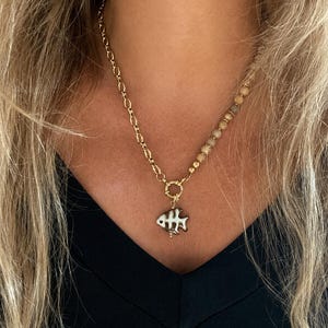 Könnte beinhalten: Eine goldfarbene Halskette mit einem gemischten Design, bestehend aus einer Kette auf der einen Seite und einer Reihe kleiner, runder, beigefarbener Perlen auf der anderen. Ein runder Goldverschluss verbindet beide Teile mit einem Fischgrätenanhänger.