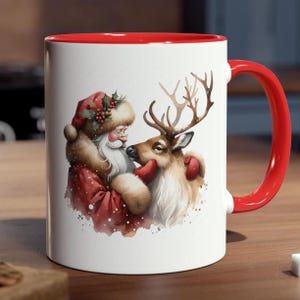 May include: A white ceramic mug with a bright red handle and interior rim. The exterior features a detailed illustration of Santa Claus in a red fur-trimmed coat and hat, gently touching the face of a brown reindeer with large antlers.