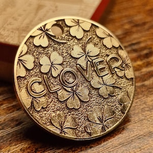 Op de afbeelding: Een gouden metalen hanger met een klavertjevier-ontwerp en het woord "CLOVER" erop gegraveerd.