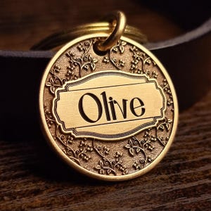 May include: A round, gold-toned metal pet identification tag featuring an embossed vine and leaf pattern surrounding a central rectangular plaque. The name Olive is engraved in a dark, serif font within the plaque, and a metal ring is attached.