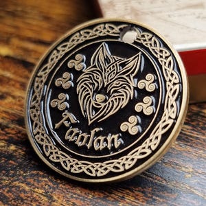 Puede incluir: Un token redondo de metal negro con un borde de nudo celta dorado. El centro presenta un diseño detallado de cabeza de lobo dorada con la palabra "Faolan" inscrita debajo.