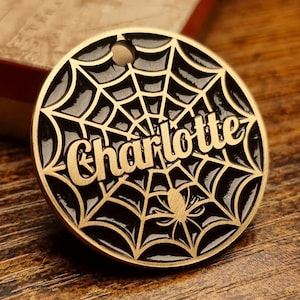 Può includere: Un'etichetta rotonda in metallo dorato con il nome "Charlotte" scritto. Presenta un disegno a ragnatela nera con un ragno al centro. C'è un piccolo foro nella parte superiore per l'attacco.