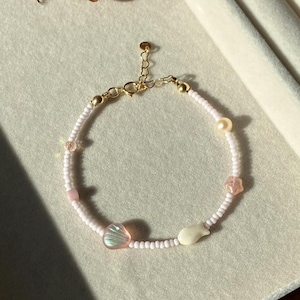 May include: A delicate bracelet featuring a strand of small white beads interspersed with pink beads and seashell charms. The bracelet includes a gold-colored clasp and a small pearl. The bracelet is designed for a dainty, feminine aesthetic.