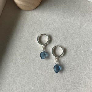 May include: A pair of silver hoop earrings with teardrop-shaped, blue gemstones. The earrings are displayed on a light gray surface, with a wooden jewelry display stand in the background. The earrings are a delicate and elegant accessory.