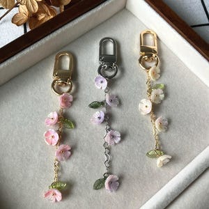 May include: Three floral keychains with gold and silver hardware. Each keychain features delicate pink and white flowers with green leaves. The keychains are displayed in a wooden tray with a white and black checkered background.