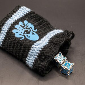 May include: Black and light blue crocheted dice bag with a light blue octopus logo. Two ornate, square dice with blue and silver accents are partially visible inside the bag. The bag is open, revealing the dice.