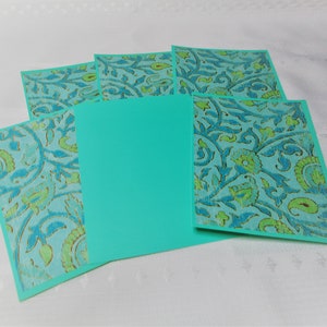 May include: A collection of turquoise notecards with a floral pattern in blue and green. The cards are arranged in a fan shape, with a solid turquoise card in the center. The background is a white, textured surface.