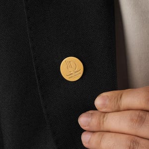 May include: A gold-colored lapel pin with a circular design, affixed to a black blazer. The pin features a stylized letter "D" and other design elements. The image shows a close-up of the pin and part of the blazer.