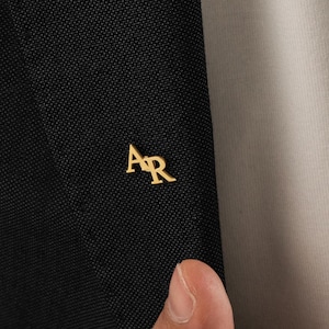 Könnte beinhalten: Nahaufnahme eines schwarzen Blazers mit einem goldfarbenen "AR"-Emblem auf dem Revers. Der Blazer ist aus einem strukturierten Stoff gefertigt und wird mit einem hellfarbigen Hemd kombiniert.