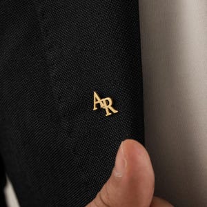 May include: A gold-colored lapel pin with the initials "AR" is affixed to a black suit jacket. The pin is small and elegant, adding a touch of sophistication to the formal attire. The jacket has a textured weave.