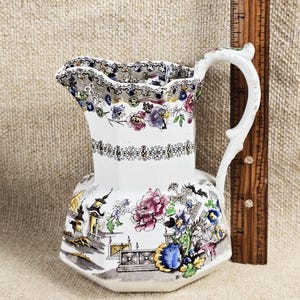 May include: A white ceramic pitcher with a floral and landscape design. The pitcher has a decorative handle and a scalloped rim. A wooden ruler is next to the pitcher. The pitcher is approximately 6 inches tall.