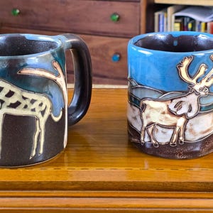 May include: Two ceramic mugs with dark brown handles and rims. The left mug features a cream-colored giraffe illustration on a dark brown background. The right mug displays a cream-colored reindeer illustration against a blue and brown landscape.
