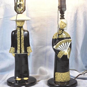 May include: A pair of ceramic table lamps shaped as figures in black and yellow patterned traditional clothing. One figure wears a wide-brimmed hat, while the other holds a white fan. Both lamps feature metal sockets with pull chains for operation.