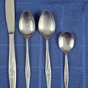 May include: A set of four stainless steel utensils including one dinner knife, two spoons of varying sizes, and one small teaspoon. Each piece features a tapered handle with a subtle embossed rose floral detail at the base of the stem.