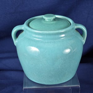May include: A turquoise ceramic pot with a lid and two handles. The pot has a rounded shape and a glossy finish. The lid has a small knob on top. The pot is sitting on a clear surface against a dark blue background.