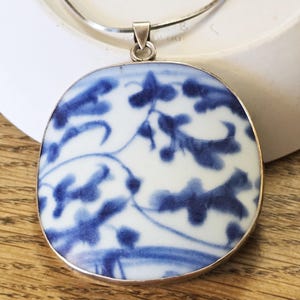 May include: A round pendant necklace with a silver chain. The pendant features a white background with a blue floral pattern and a silver border. The necklace is displayed on a wooden surface.