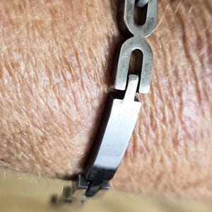 May include: A silver-toned metal link bracelet featuring a series of rectangular segments connected by U-shaped interlocking pieces. The bracelet has a polished, reflective finish and a secure clasp mechanism visible along the wrist.