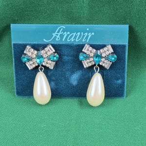 May include: A pair of earrings with a bow design, featuring clear and teal-colored crystals. Each earring has a pear-shaped, cream-colored drop. The earrings are displayed on a teal velvet card with the brand name "Aravir".