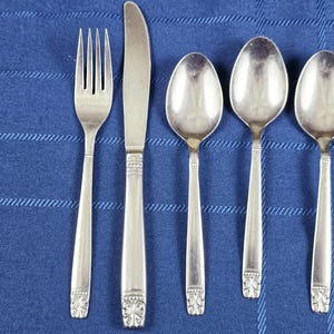 May include: A five-piece stainless steel flatware set consisting of one dinner fork, one dinner knife, and three spoons. Each piece features a polished silver finish and a decorative embossed pattern at the base of the handle, providing a uniform look.