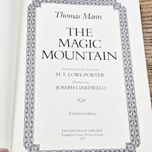 Puede incluir: Página de libro abierto con el título "THE MAGIC MOUNTAIN" de Thomas Mann. El texto está centrado en una página blanca, enmarcado por un borde decorativo. El libro es una edición limitada de The Franklin Library, publicado en 1979.