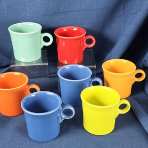 May include: A collection of six ceramic mugs in various bright colors: seafoam green, red, orange, blue, yellow, and orange. Each mug has a rounded handle and a simple, classic design, perfect for coffee or tea.