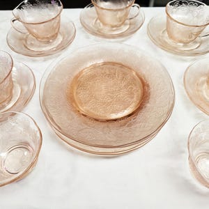 May include: A collection of vintage pink glass dishware, including teacups, saucers, and plates. The pieces feature a delicate floral pattern. The arrangement suggests a formal tea setting, perfect for a special occasion or display.