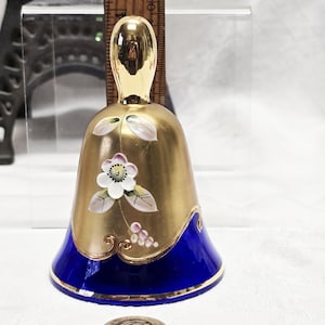 May include: A small, gold-painted glass bell with a blue base. The bell is decorated with a white flower and leaves. The bell is about 2 inches tall.