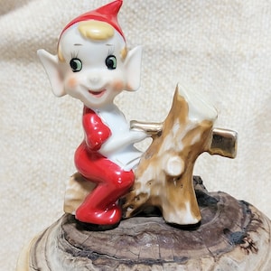 May include: A ceramic figurine of a Christmas elf wearing a red hat and red pants, sitting on a brown log.