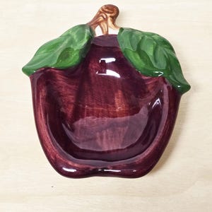 May include: A ceramic apple-shaped dish with a deep burgundy glaze. The dish features green leaves and a brown stem, adding a touch of nature-inspired decor. The glossy finish enhances the rich color and design.