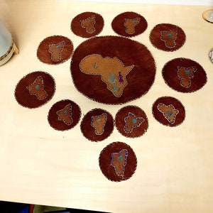 May include: A set of ten brown leather coasters with a stitched outline. Each coaster features an embroidered outline of the continent of Africa. The center coaster is larger and features a more detailed embroidery of the continent with a small figure of a person.