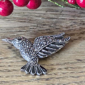 May include: A silver-toned hummingbird brooch with intricate detailing. The bird's wings are spread in mid-flight, with black enamel accents on the wings and tail. The brooch is set against a wooden background, with red berries in the background.