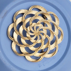 May include: A gold-toned brooch in the shape of a flower with a swirling pattern of overlapping circles. The brooch is made in England.