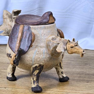 May include: A ceramic jar shaped like a horse with a speckled beige body, dark brown saddle lid, and four legs with black hooves. The horse head extends from the side, featuring sculpted ears and a mane, with dark markings on the body and legs.