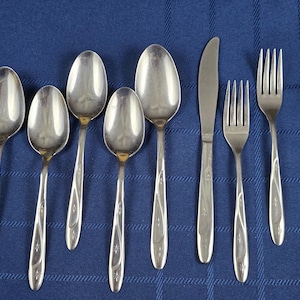 May include: A set of eight stainless steel flatware pieces including four spoons, one dinner knife, and three forks. Each piece features a polished silver finish and a slender handle decorated with a subtle, embossed swirling line and star pattern.