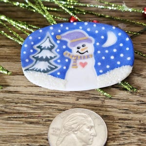 May include: Oval-shaped Christmas ornament featuring a snowman with a purple hat and scarf, a heart, and a crescent moon against a blue snowy background. A green Christmas tree is on the left. The ornament is on a wooden surface.