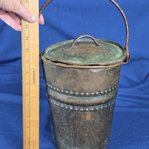 May include: Antique copper bucket with a lid and handle. The bucket is cylindrical, with a riveted design and a patinaed finish. The bucket is approximately 12 inches tall.