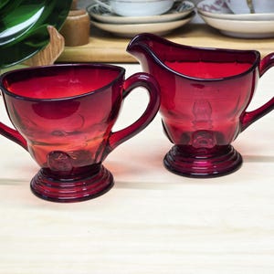 May include: A vintage ruby red glass creamer and sugar bowl set. The creamer has a single handle and the sugar bowl has two handles.