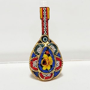 May include: A small, teardrop-shaped pendant made of colourful mosaic tiles. The pendant features a yellow flower with red and blue accents, surrounded by a green, red, and blue border. The pendant is attached to a gold chain with a red, white and yellow striped ribbon.