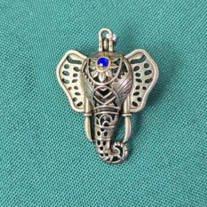 May include: A silver-toned metal pendant shaped like an elephant head with intricate cut-out filigree patterns on the ears and trunk. A small, circular, vibrant blue gemstone is set at the center of the forehead, just below a metal hanging loop.